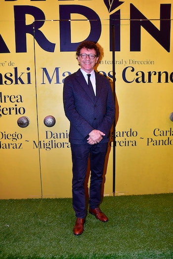 Hombre con traje y gafas sonríe, de pie sobre césped artificial. Fondo amarillo con letras grandes oscuras parcialmente visibles