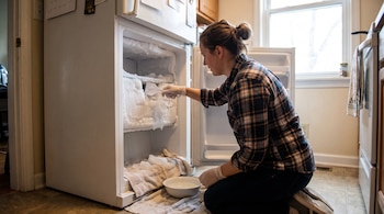Una mujer, de perfil, agachada frente a un refrigerador con el congelador abierto lleno de hielo, descongela con una pala. Hay toallas y un tazón en el suelo.