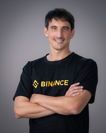 Hombre de mediana edad con cabello oscuro, bigote, sonriendo, viste camiseta negra con logo dorado de Binance y tiene los brazos cruzados sobre fondo gris