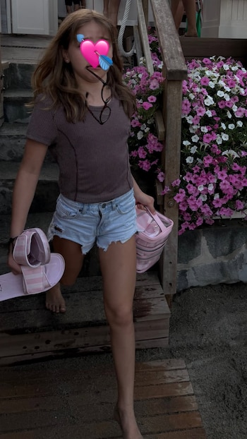 Niña con cabello castaño, camiseta de rayas y shorts de jean. Pies descalzos. Sandalias rosadas y bolso rosa. Escaleras de madera. Flores rosadas y blancas. Arena
