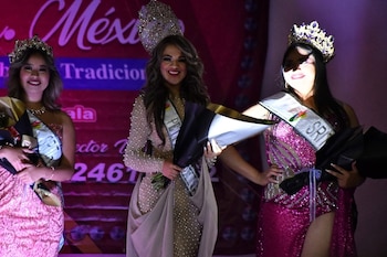 Tres mujeres con vestidos de gala, coronas y bandas sostienen ramos de flores frente a un telón de fondo oscuro
