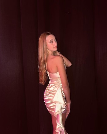 Joven con cabello largo y rubio claro, vestida con un vestido de cóctel strapless satinado color champán, posa de perfil mirando por encima del hombro derecho contra un fondo oscuro
