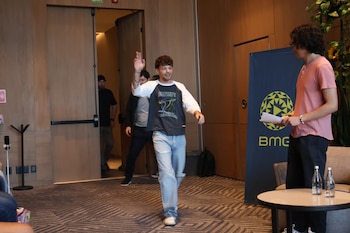 Un hombre con cabello rizado, camiseta de manga larga y jeans rasgados, saluda con la mano mientras camina en una sala. Al fondo, puertas de madera y cartel de BMG