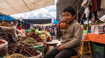 Un niño sostiene una taza humeante junto a puestos de chiles secos y hierbas en un mercado al aire libre con una montaña nevada al fondo.
