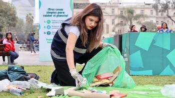 Una mujer joven con chaleco azul y guantes blancos se agacha para recoger residuos del césped y colocarlos en una bolsa verde, con un cartel ambiental de fondo