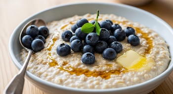 Un tazón de porridge de avena con arándanos, una hoja verde, un trozo de mantequilla y jarabe, junto a una cuchara de metal con algunos arándanos.