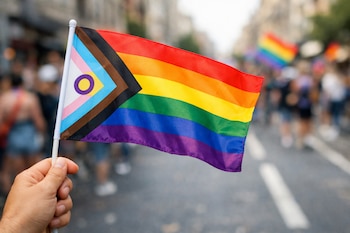 Mano sujeta bandera con franjas de arcoíris, negro, marrón, azul, rosa, blanco y círculo amarillo con anillo morado. Fondo borroso de calle con personas.