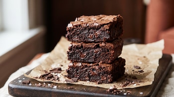 Tres trozos de brownie de chocolate apilados sobre papel, rodeados por virutas de chocolate y escamas de sal.