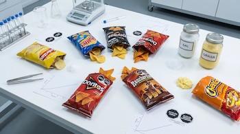 Vista cenital de mesa de laboratorio con bolsas de Sabritas, Ruffles, Doritos, Rancheritos, Cheetos abiertas, sal y grasa extraídas, balanza digital, tubos de ensayo y gráficos.