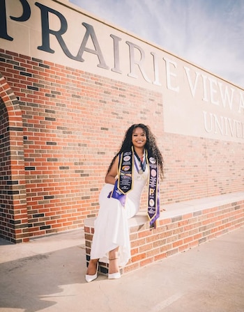 Mujer joven con pelo rizado y vestido blanco sentada en un muro de ladrillos. Viste estolas de graduación y medallas. Detrás se lee PRAIRIE VIEW A&M UNIVERSITY