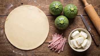Masa circular, tres zapallitos redondos, tiras de jamón cocido, cubos de queso fresco, un cuchillo y un palo de amasar sobre madera.