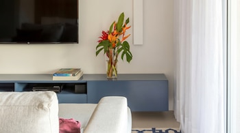 Salón moderno con televisión LG montada en la pared sobre un mueble azul flotante, un jarrón de flores tropicales y libros, junto a un sofá claro y cortinas blancas.