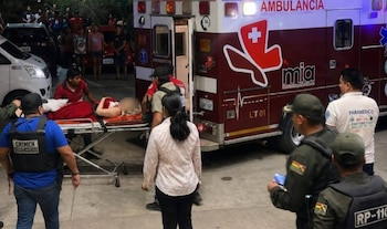 Ambulancia estacionada con puerta trasera abierta. Paramédicos y policías asisten a una persona en camilla cubierta. Varios transeúntes y vehículos en el fondo