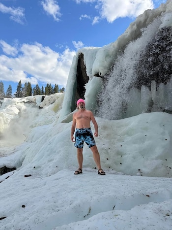 Hombre barbudo, sin camisa, con gorro rosa y pantalones cortos, de pie en la nieve frente a una cascada parcialmente congelada. Cielo azul