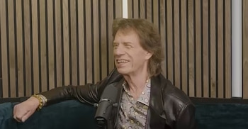 Hombre sonriendo sentado en sofá verde, con chaqueta de cuero negra, camisa estampada, reloj y pulsera. Micrófono y pared de listones de madera
