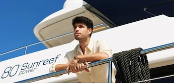 Hombre joven con camisa color crema de pie en la cubierta de un yate Sunreef blanco, con un cielo azul brillante de fondo. Sostiene una barandilla y tiene una cuerda negra enrollada a su lado