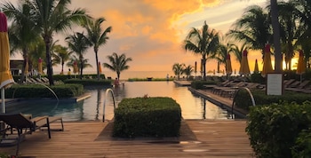 Piscinas de resort, palmeras, camastros y sombrillas bajo un cielo anaranjado al atardecer, con el mar de fondo y algunas personas en el agua