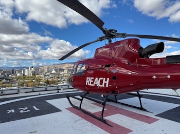 Un helicóptero rojo con la inscripción REACH está estacionado sobre la marca H de un helipuerto con una ciudad de fondo