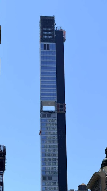 Un rascacielos moderno y alto en construcción, con fachada de cristal y una sección oscura, se eleva contra un cielo azul claro