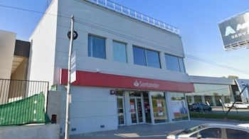 Exterior de un edificio blanco de dos pisos con un cartel rojo del Banco Santander, ventanales de vidrio y un poste en primer plano
