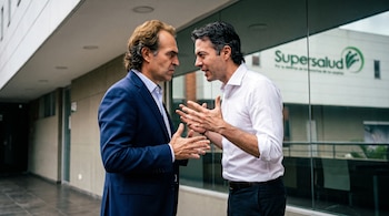 Dos hombres, Federico Gutiérrez en traje azul y Daniel Quintero en camisa blanca, se miran intensamente en un pasillo exterior. Al fondo se ve el edificio Supersalud.
