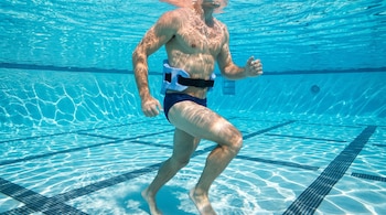 Hombre caucásico, sin camiseta, en bañador azul oscuro, con cinturón de flotación azul, trota bajo el agua en una piscina de azulejos claros y oscuros.
