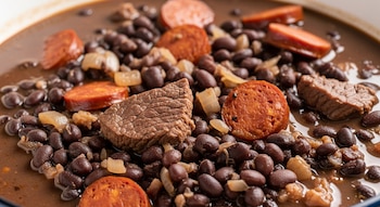 Primer plano de un guiso espeso con porotos negros, trozos de carne de res, rodajas de chorizo y cebolla picada en un caldo oscuro.