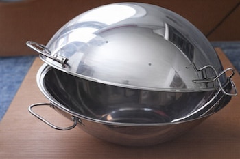 Cataplana de acero inoxidable con dos asas y tapa semiesférica abisagrada, colocada sobre una superficie de madera clara con un fondo difuminado.