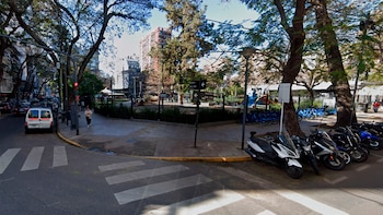 Una calle con un paso de cebra en primer plano, acera con una mujer caminando y una furgoneta blanca, junto a la entrada de la Plaza Noruega con bicicletas y árboles