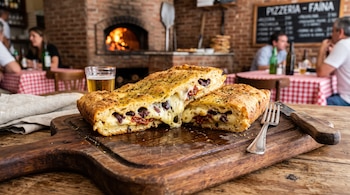 Porción de fainá cortada con relleno de queso fundido, aceitunas y tomates secos sobre una tabla de madera junto a cubiertos.