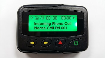 Un busca (pager) negro con pantalla verde brillante que muestra "Incoming Phone Call Please Call Ext 001". Se aprecian iconos de señal y sonido, y varios botones en la parte inferior.