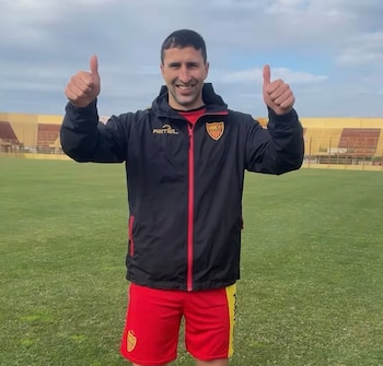 Hombre sonriente con camiseta negra y roja, pantalones cortos rojos y amarillos, levanta dos pulgares. Está en un campo de fútbol de césped verde con tribunas detrás