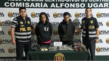 Dos agentes policiales flanquean a un hombre y una mujer detenidos frente a una mesa con varias bolsas de droga incautada y un cartel que indica 'Droga Comisada'