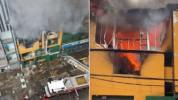 Un edificio amarillo y verde está en llamas con denso humo gris y fuego visible a través de ventanas, mientras bomberos operan un camión escalera en la calle