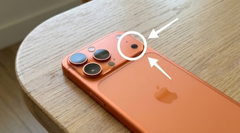 Vista superior del iPhone 17 Pro Max color coral sobre una mesa de madera clara, con un círculo y una flecha blanca señalando un pequeño sensor junto a las cámaras traseras.