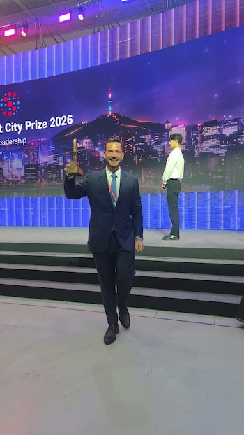 Ariel Sujarchuk, vestido de traje oscuro y corbata azul, sostiene un trofeo dorado en un escenario frente a una pantalla gigante