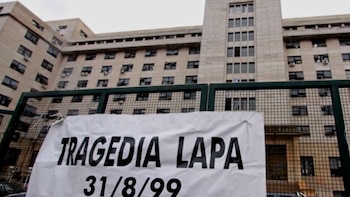 Un cartel blanco con texto negro 'TRAGEDIA LAPA 31/8/99' atado a una valla metálica oscura, con un gran edificio beige de múltiples ventanas al fondo
