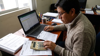 Un hombre con anteojos, suéter y polo blanco utiliza una calculadora y una laptop en una mesa de madera con documentos, taza y portalápices.