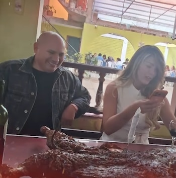 Hombre calvo y mujer joven sentados a una mesa. Hay un gran plato de carne asada, una botella de vino y un teléfono móvil en la mesa