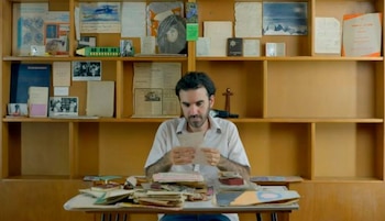 Hombre con barba sentado en un escritorio de madera, examinando un papel. El escritorio tiene pilas de documentos y libros. Detrás, una estantería llena de archivos y fotos