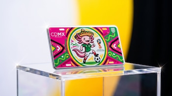 Primer plano de una placa vehicular conmemorativa de la CDMX para el Mundial 2026, con un ajolote futbolista, sobre un pedestal acrílico, fondo blanco, amarillo y negro.