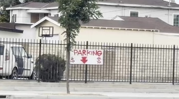 Un cartel blanco con la palabra 'PARKING' en rojo y una flecha hacia abajo con símbolos de dólar, colgado en una valla negra, con casas y un coche blanco detrás