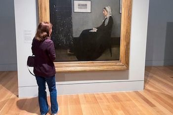 Una persona de espaldas con chaqueta morada y jeans observa un gran cuadro enmarcado en oro que representa a una anciana sentada de perfil