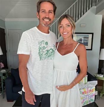 Una pareja sonriente posa en el interior de su casa. La mujer, con un vestido blanco, se toca el vientre embarazado. El hombre lleva una camiseta blanca