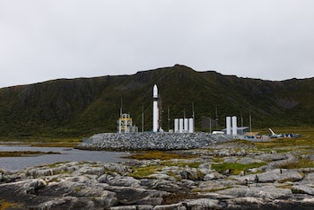 Cohete blanco en posición vertical junto a varios tanques cilíndricos, sobre una plataforma de rocas con montañas y vegetación al fondo