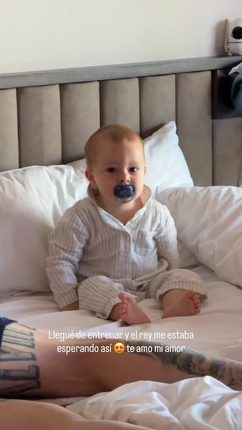Un bebé vestido con pijama de rayas y chupete azul permanece sentado en una cama con sábanas y almohadas blancas