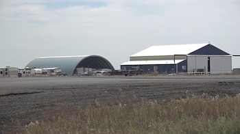 Una vista de un aeródromo con dos grandes hangares, uno azul y blanco y otro curvado, junto a varios vehículos recreativos estacionados bajo un cielo nublado