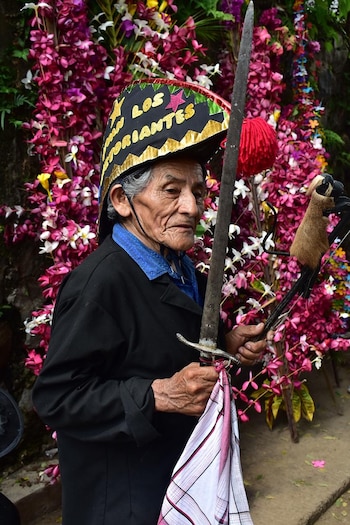 Retrato de un hombre mayor con sombrero negro y dorado, chaqueta oscura, sosteniendo una espada y un objeto con piel, frente a una pared densa de flores rosadas, moradas y blancas