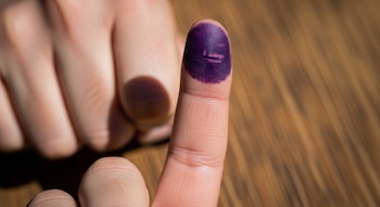 Un dedo índice con tinta morada indeleble, junto a una mano difuminada y un fondo de madera granulado.