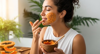 Mujer con cabello oscuro aplica mascarilla facial de papaya en su mejilla y boca con los dedos, sosteniendo un cuenco. Rodajas de papaya en la mesa.